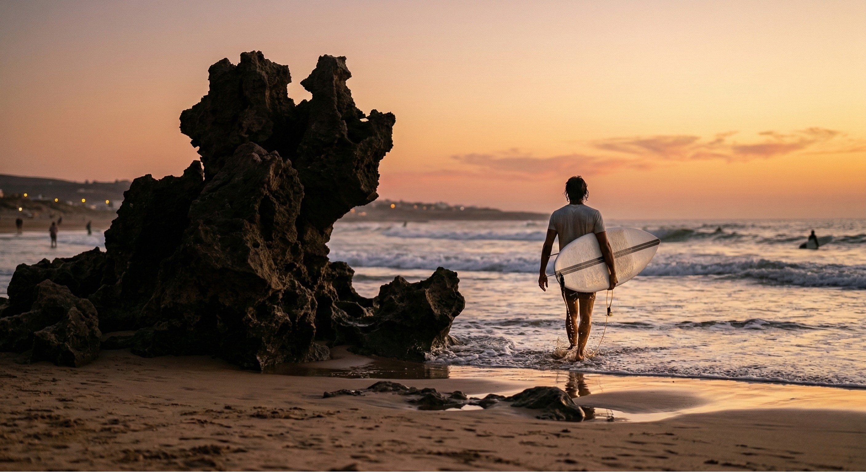 A surfer carrying a surfboard on the beach at Devil's Rock in Tamraght during golden hour, with the sun reflecting off the ocean waves